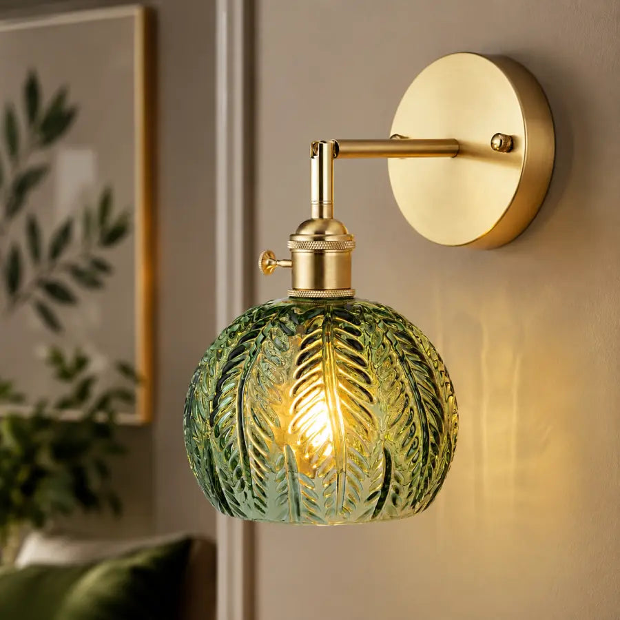 A wall-mounted light fixture with a round green textured glass shade and a gold-finished wall plate and arm, shown lit against a beige wall in an interior setting.