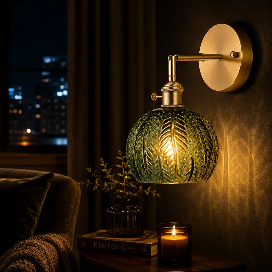 A wall-mounted sconce with a round gold backplate, brass-toned arm, and a textured green glass shade glowing warm yellow, shown lit in a dimly lit room with a night city window, candle, and book in the foreground.