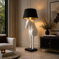 A modern floor lamp shaped like a white horse standing on a black base, with a black lampshade and warm gold interior, placed in a styled living room beside a sofa and sideboard.